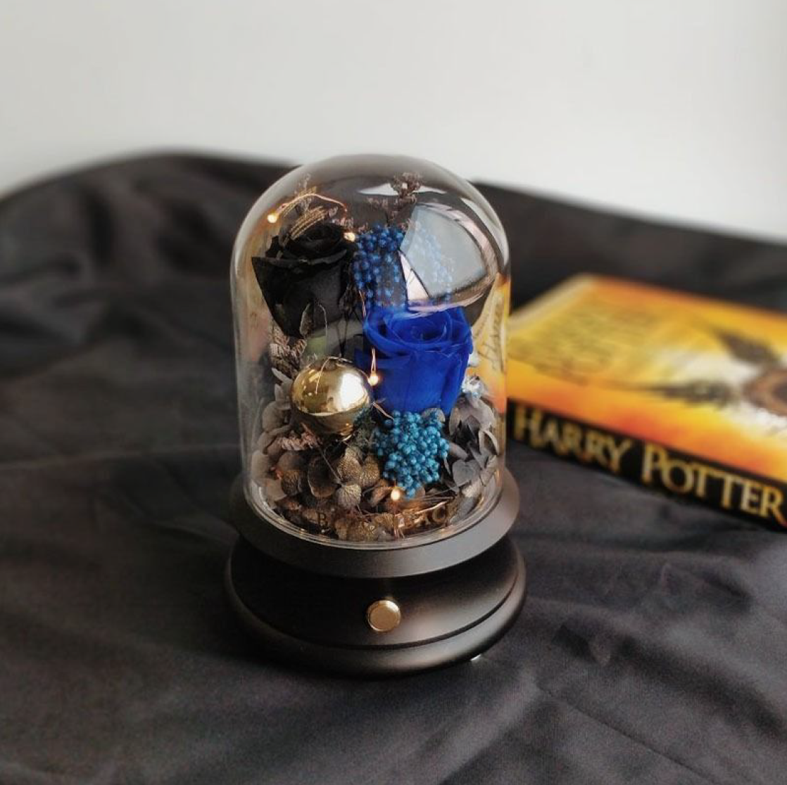 Blue and black rose in glass dome with the bluetooth speaker embedded inside and black base. Behind is harry potter book in the background.
